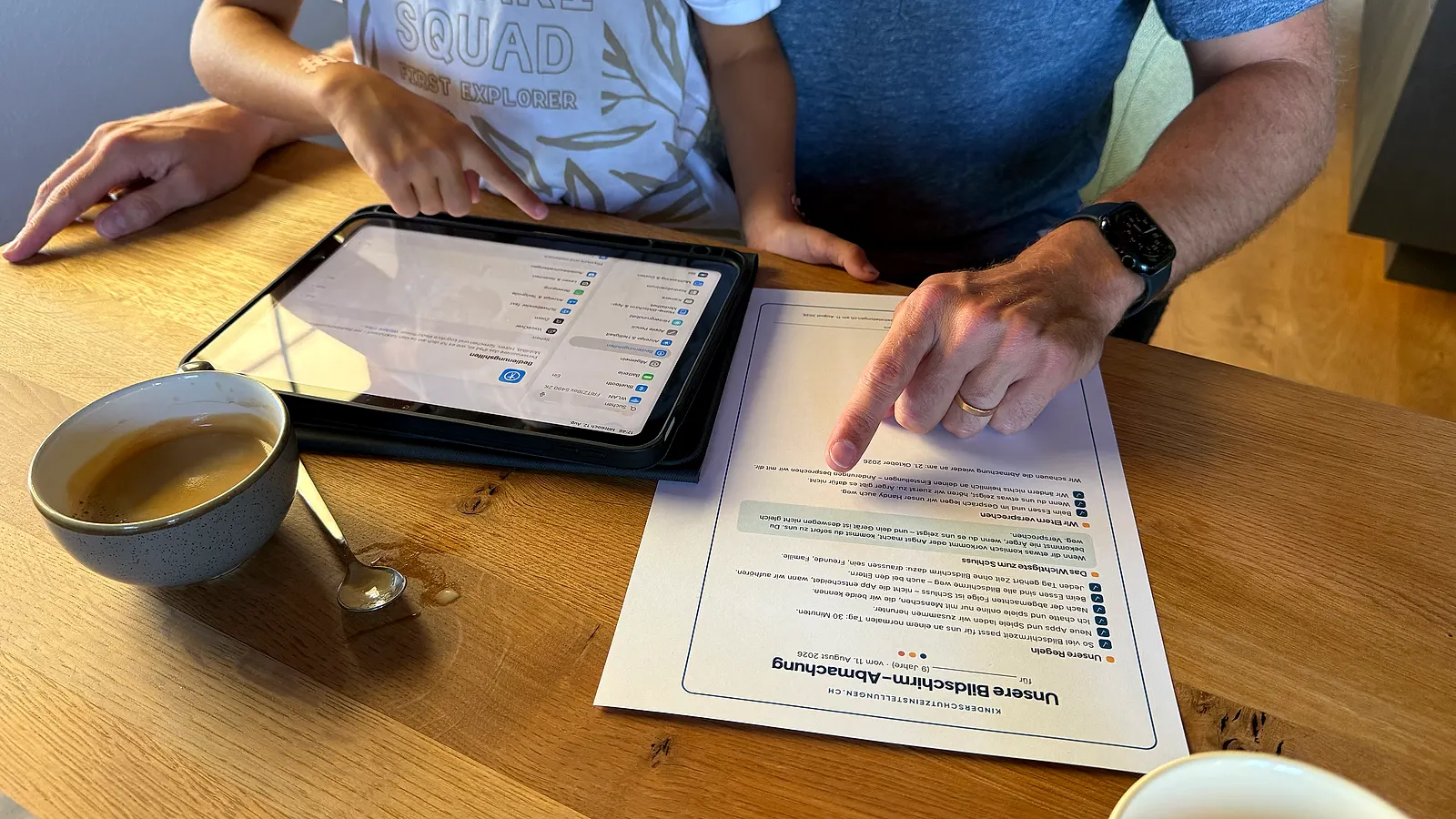 Vater und Kind sitzen am Esstisch: Der Vater zeigt auf die ausgedruckte Bildschirm-Abmachung, daneben liegt ein Tablet mit den geöffneten Einstellungen